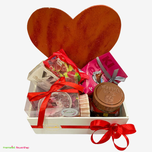 Vista superior de caja de madera con café Juan Valdez, chocolates, jabones naturales y vela aromática, sobre fondo de corazón rojo con glitter.