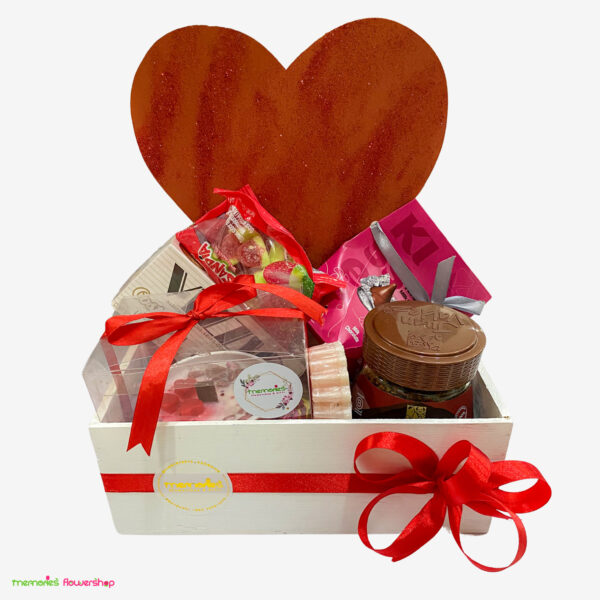 Vista frontal de caja de madera con café Juan Valdez, chocolates, jabones naturales y vela aromática, sobre fondo de corazón rojo con glitter.